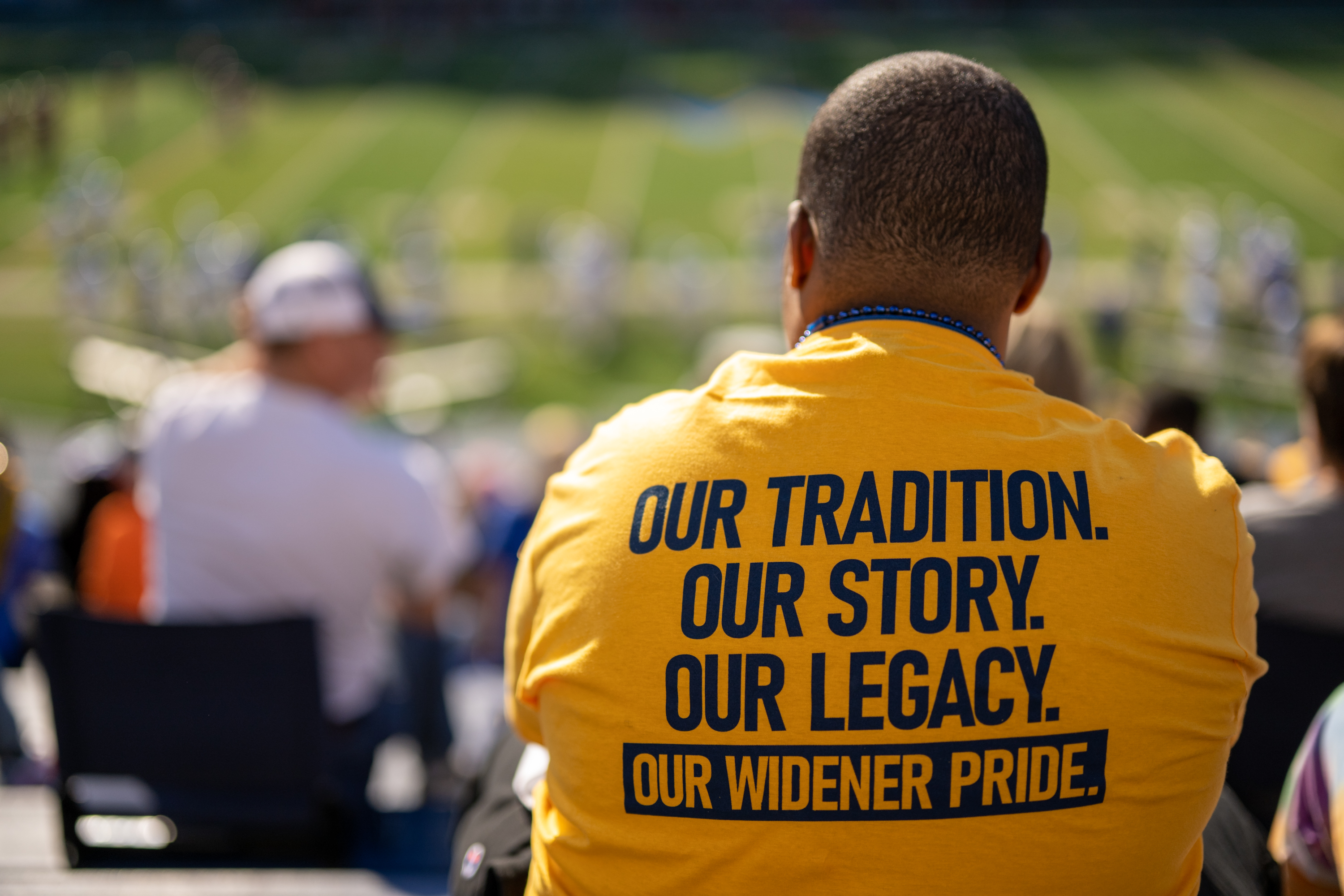 An alumnus sits in the stands and the back of his shirt reads "our tradition, our story, our legacy."